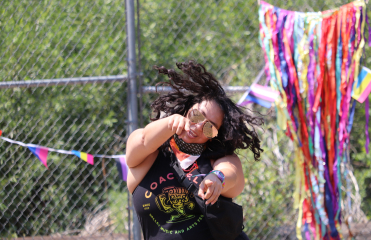 Happy woman dancing and pointing at the camera with rainbow streamers hanging from a chainlink fence behind her