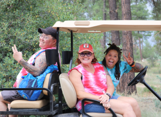 Three people sitting on a golf cart smiling and gesturing peace signs