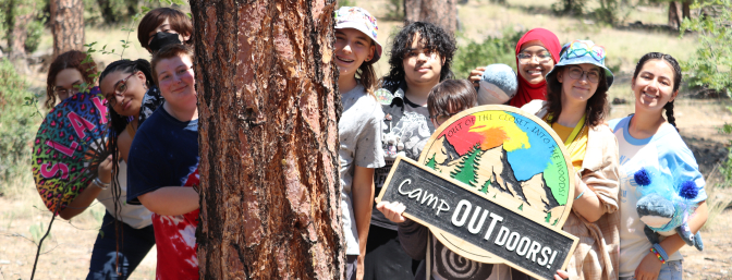 Camp Outdoor attendees standing next to a tall pine tree holding a wooden Camp Outdoors sign