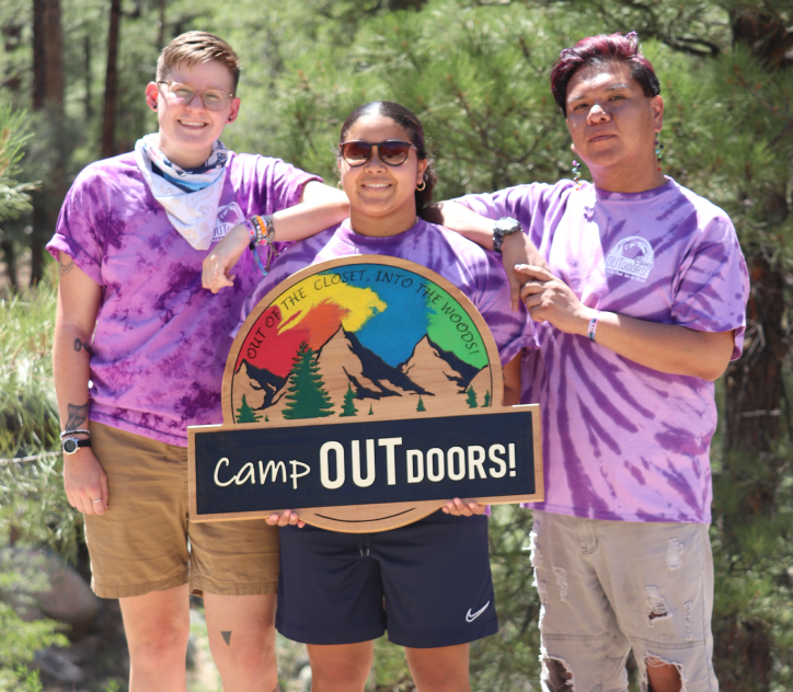 Camp Outdoor volunteers standing in front of pine trees, wearing matching white and purple tie-dyed tank tops holding a wooden Camp Outdoors sign