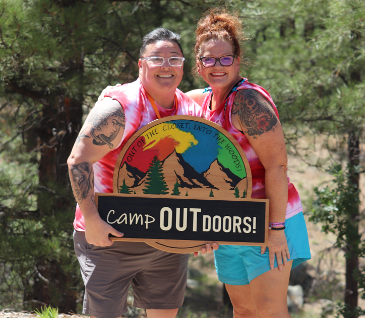 Camp Outdoor volunteers standing in front of pine trees, wearing matching white and pink tie-dyed tank tops holding a wooden Camp Outdoors sign