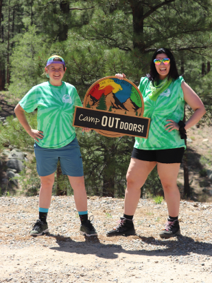 Two women in green and white tie-dyed shirts standing in the woods holding a wooden Camp Outdoors sign