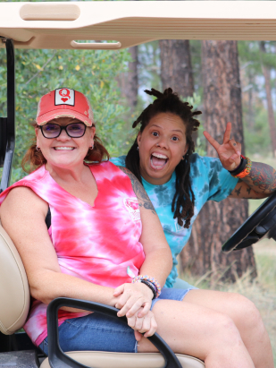 Two women sitting on a golf cart in the woods, smiling and gesturing a peace sign