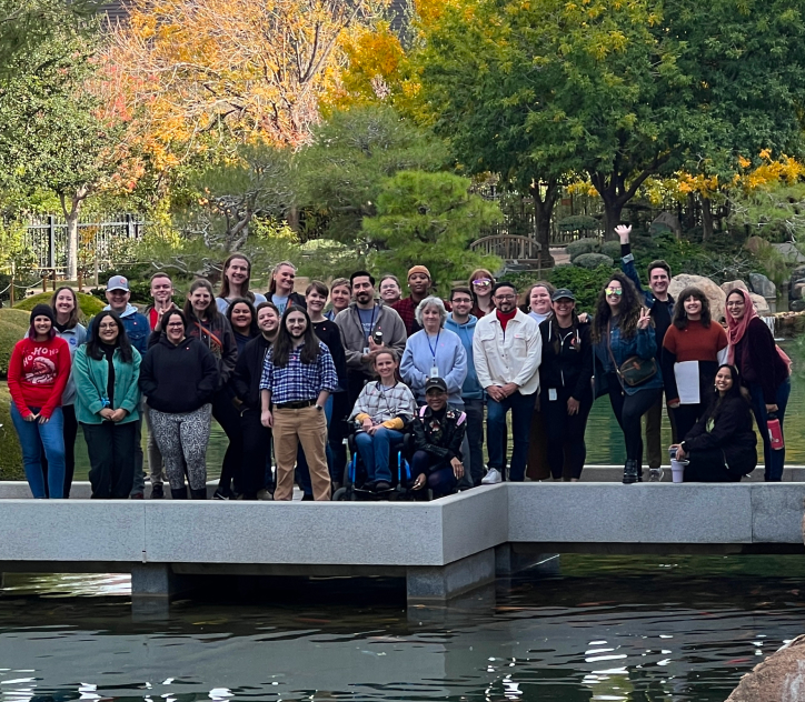 Team of people standing outside with lush green trees in the back and a body of water in front