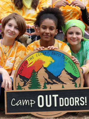 Camp Outdoor youth sitting, wearing matching white and yellow tie-dyed t-shirts holding a wooden Camp Outdoors sign