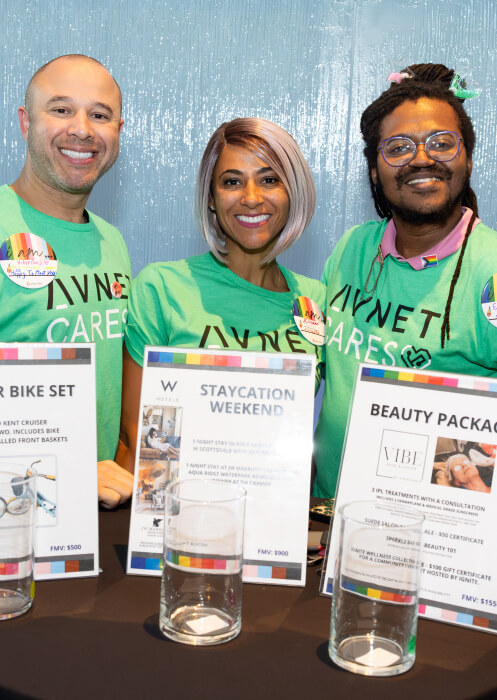 Three adults dressed in Avnet Cares t-shirts standing in front of auction items for an LGBTQ fundraising event