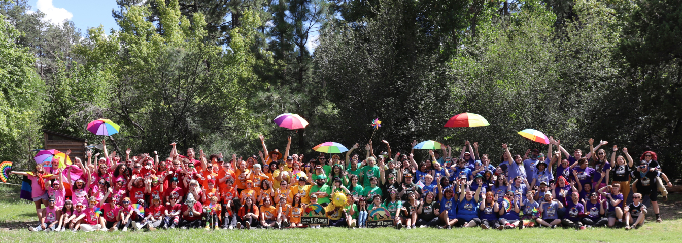 Very large group of people all dressed in Camp Outdoors t-shirts grouped by color. Some people have rainbow umbrella. There are two wooden Camp Outdoors signs in front of the group.