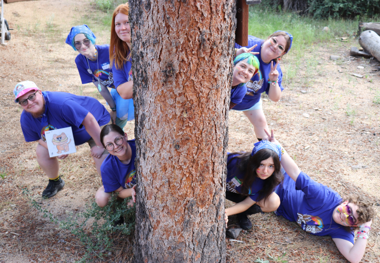 Group of people wearing matching purple Camp Outdoors t-shirts while posing behind a thick pine tree trunk