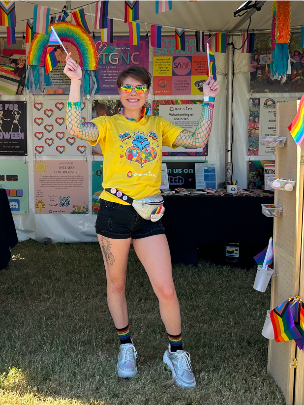 LGBTQ female wearing a brightly colored shirt, holding LGBTQ flags and standing at an outdoor booth decorated with brightly colored posters