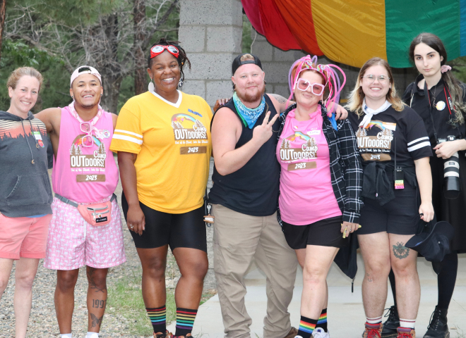 Group of Camp Outdoors attendees and volunteers standing in front of a rainbow flag