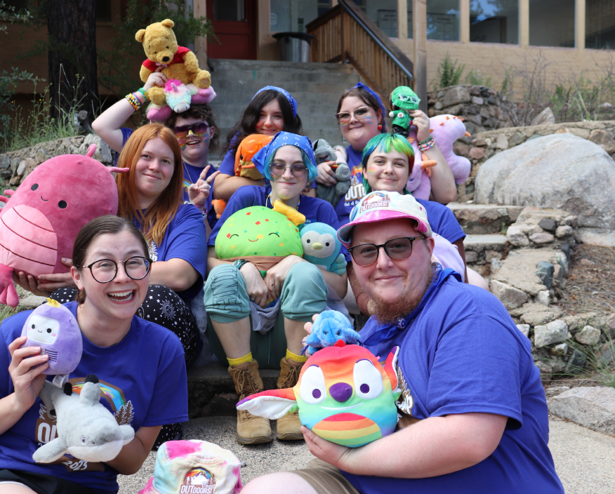 LGBTQ youth, teens and young adults showing off their Squishmallows and stuffed animals. All of them are wearing matching purple Camp Outdoors t-shirts.