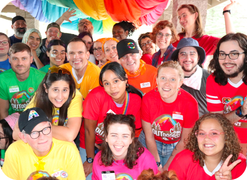 Group of camp outdoors attendees all wearing different colored Camp Outdoors shirts, holding their arms up and smiling