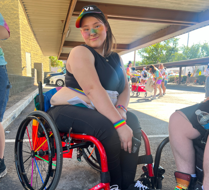 LGBTQ female in a wheelchair wearing rainbow bracelets, face paint and a flag