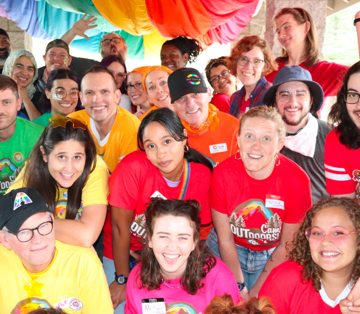 Group of camp outdoors attendees all wearing different colored Camp Outdoors shirts, holding their arms up and smiling