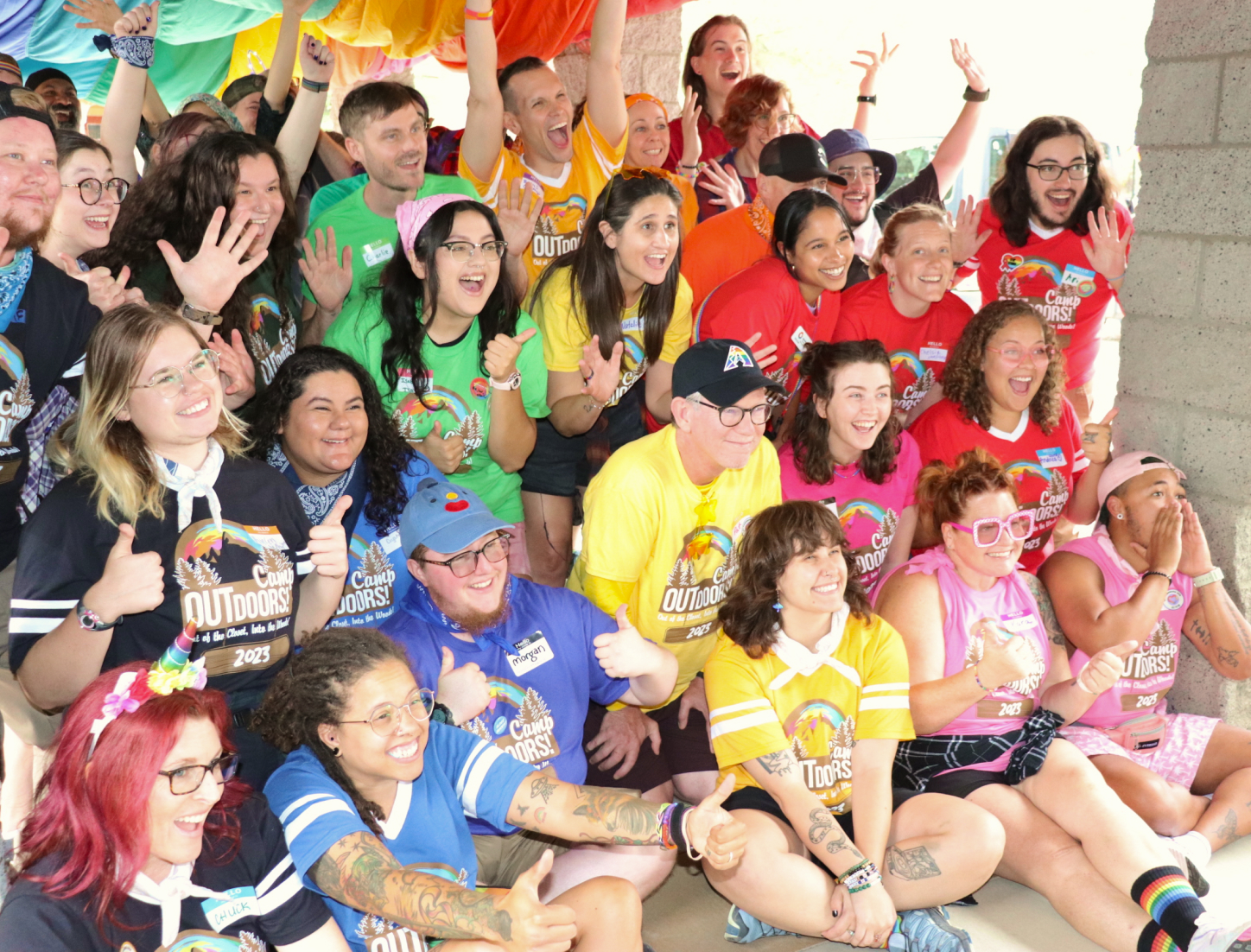 Group of camp outdoors attendees all wearing different colored Camp Outdoors shirts, holding their arms up and smiling