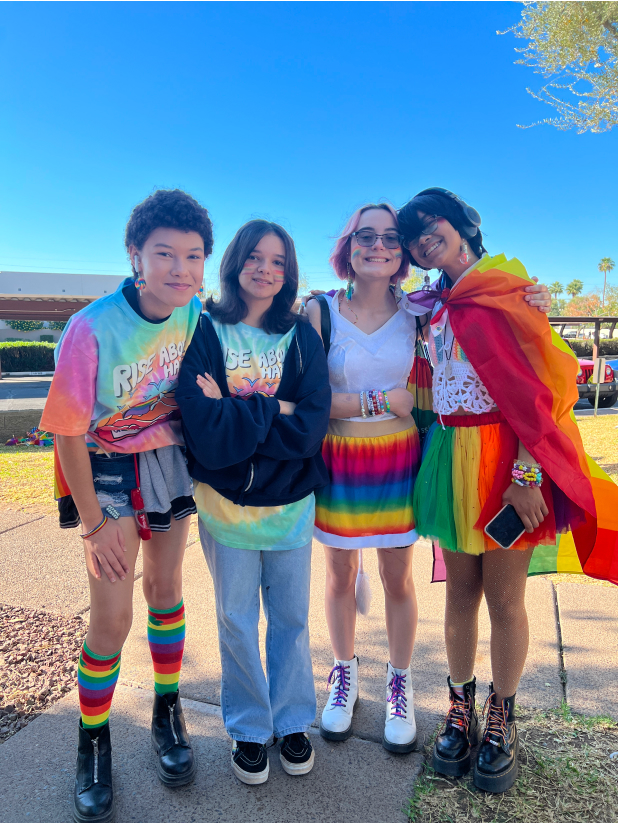 Group of four LGBTQ youth wearing rainbow clothing, standing outside and smiling. Some have their arms wrapped around one another.