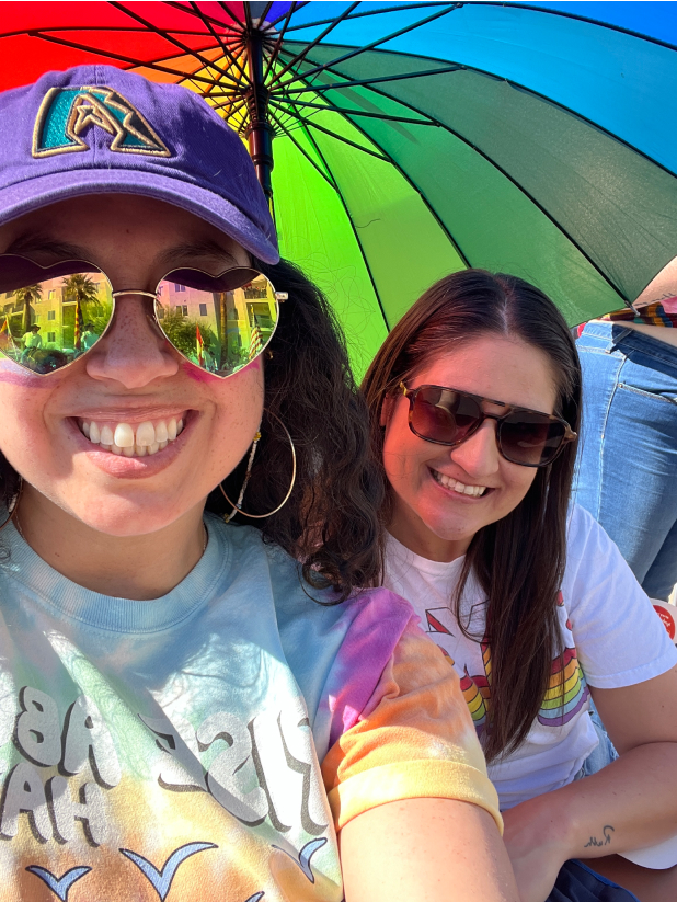 Two females wearing sunglasses and smiling under a rainbow umbrella.