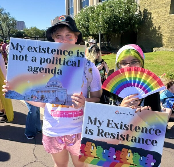 Two young adults at a pride parade holding signs that say "My existence is not a political agenda" and "My existence is resistance" and holding a rainbow fan covering their faces.